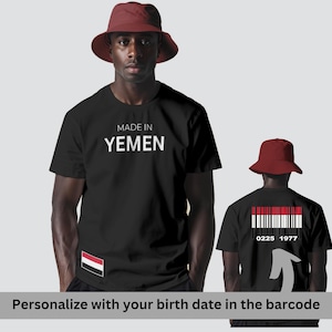 Puede incluir: Camiseta negra con texto blanco "MADE IN YEMEN". La camiseta presenta la bandera yemení en la manga y un diseño de código de barras en la espalda con los números "0225 1977". Se lleva un sombrero de pescador rojo.