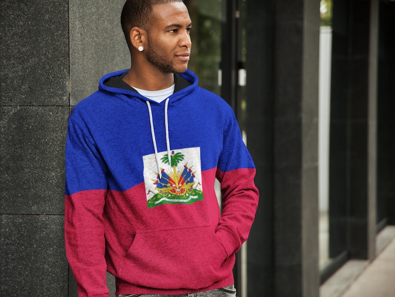 May include: A blue and red hoodie with the flag of Haiti printed on the front. The flag features a coat of arms with a palm tree, a cannon, and a drum.
