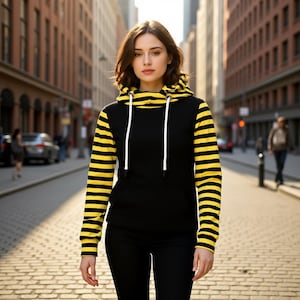 May include: Black hoodie with yellow striped sleeves and hood. The hoodie features a front pocket and white drawstrings. The person is wearing black trousers. The background is a city street with brick pavements and tall buildings.