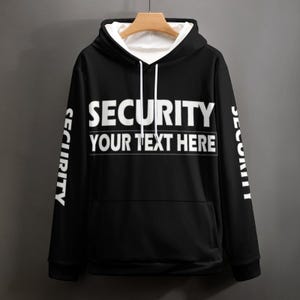 Può includere: Felpa con cappuccio nera con testo bianco "SECURITY" e "YOUR TEXT HERE" sul petto. La parola "SECURITY" è stampata anche verticalmente sulle maniche. La felpa ha un interno bianco e un cappuccio con coulisse.