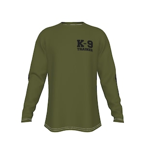 May include: Olive green long-sleeve shirt with the text "K-9 TRAINER" printed in black on the upper chest. The shirt has white stitching along the hem and cuffs. The sleeves have a black design on the upper arm.