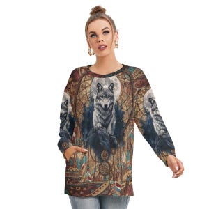 May include: A long-sleeved sweatshirt featuring a detailed wolf design. The artwork includes a wolf under a full moon, surrounded by dreamcatcher and feather motifs. The color palette is dominated by earthy tones with accents of blue and red.