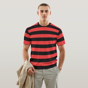Könnte beinhalten: Ein kurzärmeliges T-Shirt mit roten und schwarzen Streifen. Die Person trägt eine hellgraue Hose und eine beige Jacke. Der Hintergrund ist hellgrau.