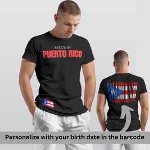 Peut inclure: T-shirt noir avec l'inscription "MADE IN PUERTO RICO" en rouge. Il présente un petit drapeau portoricain à l'avant et un motif de code-barres du drapeau au dos, avec "1993 0407" imprimé en dessous. Le t-shirt est porté par une personne.