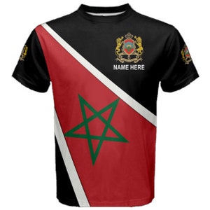 Può includere: Una maglietta nera con un design diagonale che presenta la bandiera marocchina. La bandiera è rossa con una stella verde a cinque punte e una striscia bianca. La metà superiore della maglietta è nera con uno stemma dorato e il testo "NAME HERE".