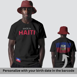 Può includere: T-shirt nero con la scritta "MADE IN HAITI" in rosso. La parte anteriore della maglietta presenta una piccola bandiera haitiana. La parte posteriore della maglietta presenta un design a codice a barre con la bandiera haitiana e la scritta "1993 0407". La persona indossa un cappello da pescatore rosso.