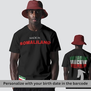 Könnte beinhalten: Schwarzes T-Shirt mit dem roten Text "MADE IN SOMALILAND". Das Shirt zeigt ein Somaliland-Flaggen-Design und einen Barcode mit dem Text "1993 0407". Die Person trägt einen roten Fischerhut.