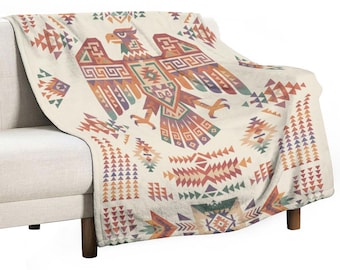 May include: A cream-coloured throw blanket draped over a sofa. The blanket features a large, colourful eagle design with geometric patterns in shades of orange, red, green, and purple. The blanket has a soft, plush texture.