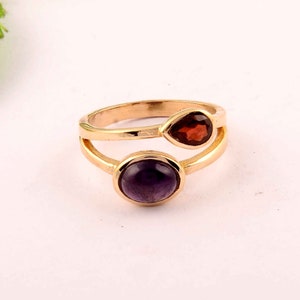 Pode incluir: Um anel de ouro com duas pedras preciosas. A pedra maior é uma ametista redonda e roxa. A pedra menor é um granate vermelho em forma de lágrima.