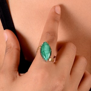 Puede incluir: Un anillo dorado con una piedra de malaquita verde vibrante en forma de marquesa. El anillo tiene una banda detallada y texturizada y un engaste plateado. El anillo se muestra en un dedo.
