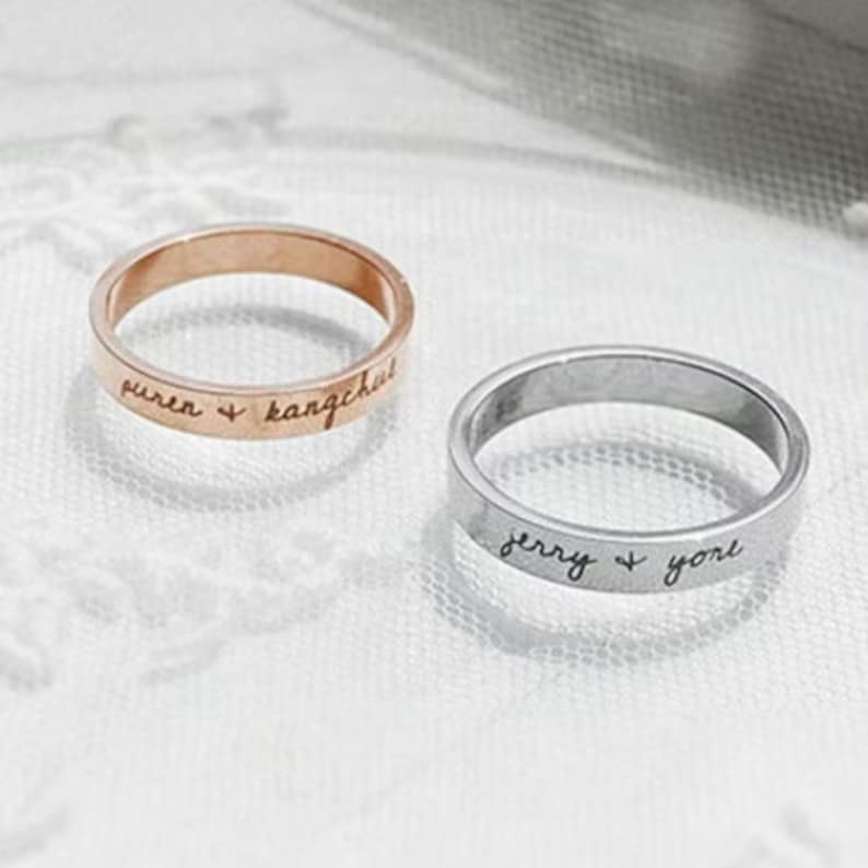 May include: Two simple silver and rose gold bands with engraved names and a heart symbol. The bands are on a white surface.