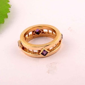 Könnte beinhalten: Ein Goldring mit geometrischem Design und violetten Edelsteinen. Der Ring hat ein käfigartiges Design mit offenen Stellen.