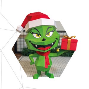 Puede incluir: Una figura de papel del Grinch verde, con gorro de Papá Noel y bufanda roja, sosteniendo una caja de regalo roja. La figura está hecha de formas geométricas. Se muestra el texto "GRINCH" y las dimensiones (27 cm x 25 cm x 15 cm). Logo de Paperking.