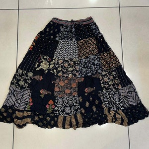 Op de afbeelding: Een zwarte maxi-rok met een patchwork-ontwerp met verschillende bloemen- en geometrische patronen in tinten bruin, crème en roze.