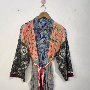 May include: A colorful, patterned robe with a tie belt. The robe features a mix of floral and paisley designs in shades of pink, blue, gray, and teal. The sleeves are long and wide, and the robe has pockets. The robe is hanging on a wooden hanger.
