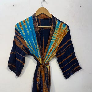Könnte beinhalten: Ein Robe mit Batikmuster in Blau-, Braun- und Gelbtönen hängt an einem Holzbügel. Die Robe hat ein Wickeldesign mit passendem Bindegürtel. Der Stoff hat ein strukturiertes Aussehen und die Ärmel sind dreiviertellang.