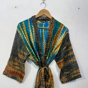 May include: A tie-dye patterned robe in shades of blue, gold, and brown hangs on a wooden hanger. The robe features wide sleeves and a matching tie belt. The fabric has a textured appearance, with a mix of vertical and horizontal patterns.
