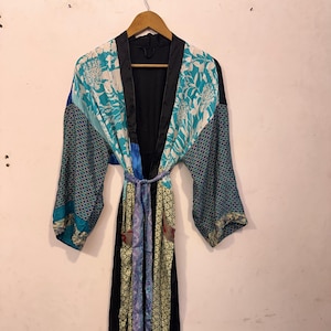 Puede incluir: Una bata larga y abierta con un diseño colorido y estampado. La bata presenta un interior y ribete negros, con mangas y paneles en tonos turquesa, azul y amarillo. Un cinturón a juego ciñe la cintura. La bata está colgada en una percha de madera.