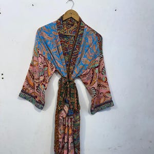 May include: A colorful paisley print robe with long sleeves, hanging on a wooden hanger. The robe features a mix of blue, pink, orange, and green hues. The robe is tied at the waist with a matching fabric belt.