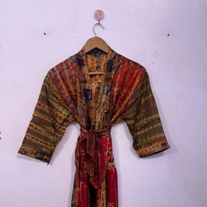 May include: A colorful, patterned robe with a tie belt, hanging on a wooden hanger. The robe features a mix of red, brown, and green hues in a patchwork design. The sleeves are three-quarter length.
