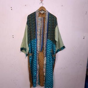 Può includere: Lunga vestaglia kimono fantasia con un mix di colori e texture. La vestaglia presenta motivi floreali e geometrici nei toni del verde acqua, verde e oro. Ha maniche ampie e tasche, ed è appesa a una gruccia di legno.