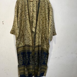 May include: A long, open-front kimono robe with a floral pattern in shades of tan, olive green, and navy blue. The robe is hanging on a wooden hanger against a white wall. The bottom section features a navy blue border.