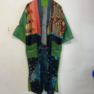 May include: A patchwork kimono robe with a variety of patterns and colors. The robe features green sleeves and pockets, with panels of red, brown, and blue floral designs. The robe is hanging on a wooden hanger.