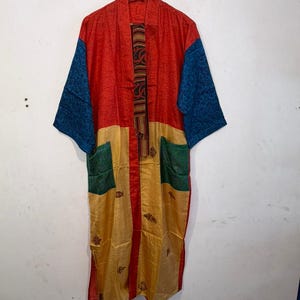 May include: A long-sleeved robe featuring a color-blocked design with a red upper section, blue sleeves, and a gold lower section. It includes two green patch pockets, a red front placket, and a thin, striped tie belt hanging from the collar.