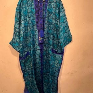 Pode incluir: Um quimono longo e fluido em tons de azul-petróleo e azul, com um design estampado. O quimono apresenta uma borda roxa contrastante ao longo das bordas e bolsos. Está pendurado em um cabide de madeira.