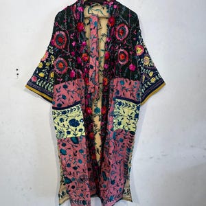 Könnte beinhalten: Ein langer, offen geschnittener Kimono mit Patchwork-Design. Der Kimono hat gestickte Blumenmuster in Rosa, Gelb und Blau auf einem dunkelgrünen Hintergrund. Die Ärmel haben einen gelben Besatz. Das Kleidungsstück hängt an einem Holzbügel.