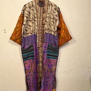 May include: A long, open-front kimono robe with a patchwork design. The robe features a mix of colors and patterns, including paisley, floral, and geometric prints. The sleeves are a burnt orange color, and the body of the robe is a mix of cream, purple, and patterned fabrics.
