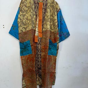 May include: A patchwork kimono robe with a variety of patterns and colors. The robe features a beige floral print, blue sleeves, and paisley and geometric designs in shades of orange, brown, and teal. It has two front pockets and an orange accent down the center.