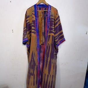 May include: A long, open-front kimono robe with a tie-dye pattern in shades of purple, orange, and blue. The robe is hanging on a wooden hanger against a white wall. The sleeves are wide and the fabric appears lightweight.