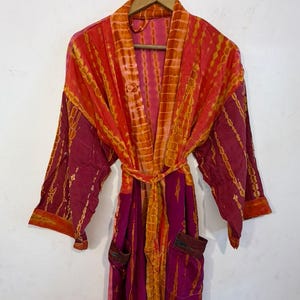 May include: A long-sleeved robe featuring a vibrant tie-dye pattern in shades of magenta, orange, and yellow. The garment includes a matching tie belt at the waist, two front pockets, and a wide collar, all hanging on a wooden hanger.