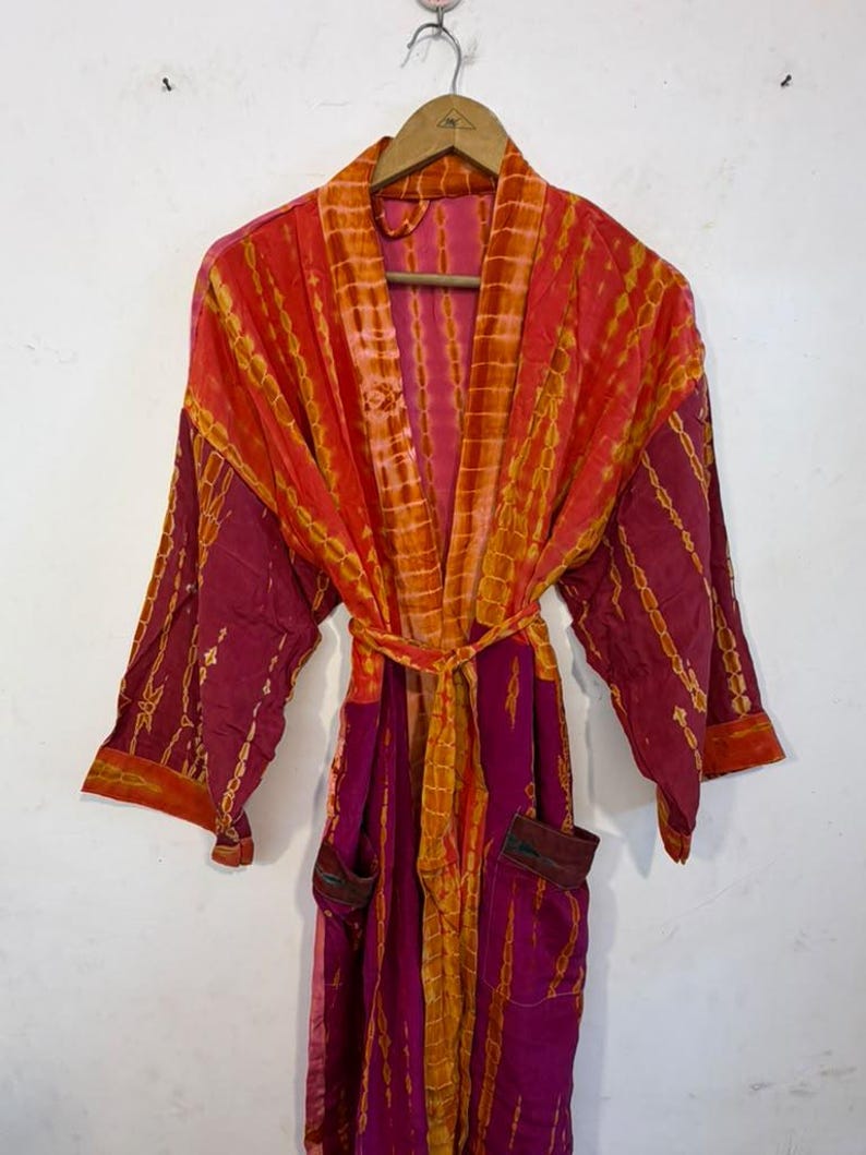 May include: A long-sleeved robe featuring a vibrant tie-dye pattern in shades of magenta, orange, and yellow. The garment includes a matching tie belt at the waist, two front pockets, and a wide collar, all hanging on a wooden hanger.
