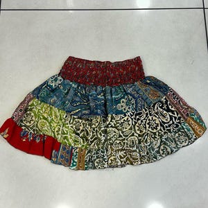 May include: A patchwork mini skirt with a red elasticated waistband and a ruffled hem. The skirt features patterned fabrics in blue, green, red, and white. The skirt is designed for casual wear.