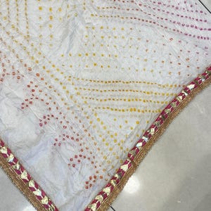 May include: White fabric with a dotted pattern in yellow, orange, and pink. The edge is trimmed with a gold border and pink accents. The fabric has a textured appearance.