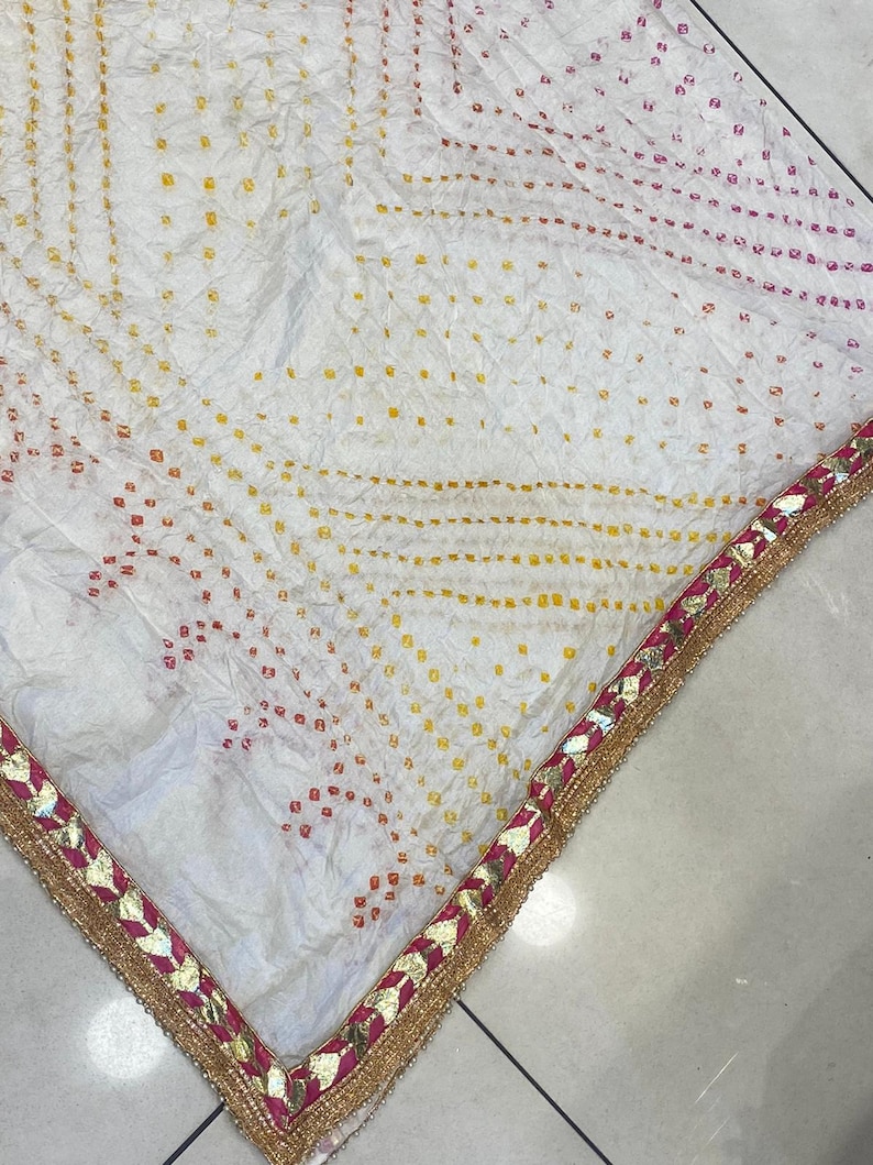 May include: White fabric with a dotted pattern in yellow, orange, and pink. The edge is trimmed with a gold border and pink accents. The fabric has a textured appearance.