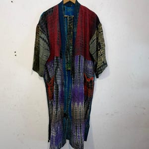 May include: A long-sleeved kimono-style robe featuring a patchwork design with tie-dye patterns in shades of deep red, purple, blue, olive green, and black. The garment has an open front, a wide collar, and two front pockets on the lower panels.