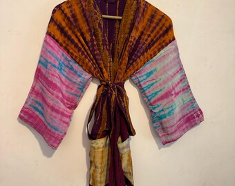 May include: A colourful tie-dye kimono, featuring orange, purple, pink, and blue hues. The robe has wide sleeves and a tie waist. The fabric looks lightweight and flowing, suitable for layering. The garment is hung on a wooden hanger.