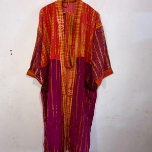 May include: A long-sleeved kimono-style robe featuring a color-blocked design in vibrant shades of magenta and orange. The garment displays a vertical tie-dye pattern throughout, with a wide front placket and an open-front silhouette.