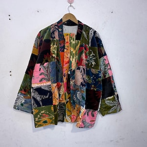 May include: A multi-coloured patchwork jacket featuring floral and geometric designs in green, pink, orange, and blue. The jacket is suspended on a wooden hanger against a white wall.