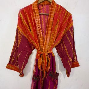 May include: A long-sleeved robe featuring a vibrant tie-dye pattern in shades of magenta, orange, and yellow. The garment includes a matching tie belt at the waist, wide sleeves, and two front pockets, all displayed on a wooden hanger.
