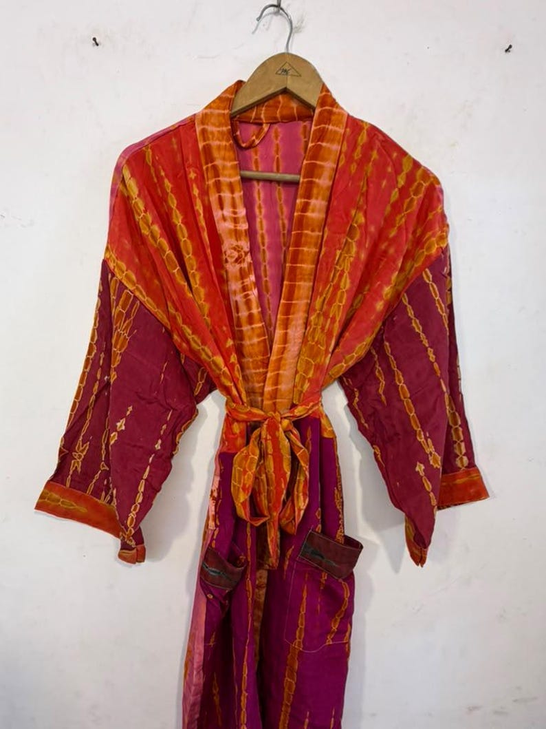 May include: A long-sleeved robe featuring a vibrant tie-dye pattern in shades of magenta, orange, and yellow. The garment includes a matching tie belt at the waist, wide sleeves, and two front pockets, all displayed on a wooden hanger.