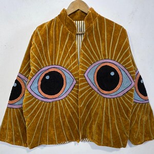 May include: Mustard-colored velvet jacket with embroidered eye designs. The jacket features large, stylized eyes in shades of black, blue, orange, and purple, radiating sunburst patterns. The jacket is hanging on a wooden hanger.