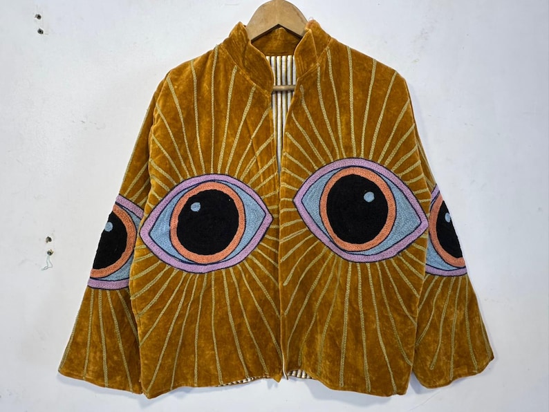 May include: Mustard-colored velvet jacket with embroidered eye designs. The jacket features large, stylized eyes in shades of black, blue, orange, and purple, radiating sunburst patterns. The jacket is hanging on a wooden hanger.