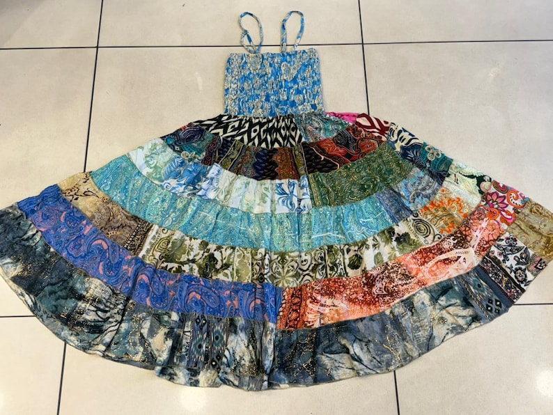 Puede incluir: Un vestido patchwork con un corpi&ntilde;o azul fruncido y tirantes finos. La falda presenta una variedad de parches de tela coloridos en tonos azules, verdes, naranjas y rosas. El vestido est&aacute; sobre una superficie embaldosada.