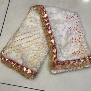 May include: White textile with yellow and orange dot patterns. The edges are trimmed with gold, pink, and gold leaf-shaped accents. A small orange tassel hangs from one end. This textile is likely a scarf or shawl.