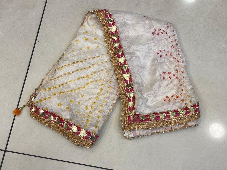 May include: White textile with yellow and orange dot patterns. The edges are trimmed with gold, pink, and gold leaf-shaped accents. A small orange tassel hangs from one end. This textile is likely a scarf or shawl.