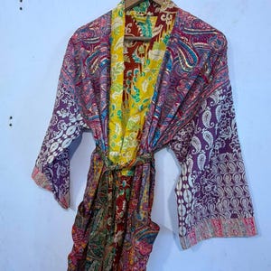 Może przedstawiać: Szlafrok typu kimono z długimi rękawami, ozdobiony żywym wzorem patchworkowym z r&oacute;żnymi motywami kwiatowymi i paisley w odcieniach ż&oacute;łci, głębokiej czerwieni, fioletu i r&oacute;żu. Odzież posiada pasujący pasek z tkaniny oraz jaskrawoż&oacute;łty kołnierz wewnętrzny.